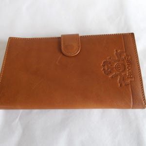 May include: A rectangular, brown leather wallet with a snap closure. The wallet features a textured border on the right side and an embossed design with the name "Brocchieri". The wallet is closed with a small, rounded tab.