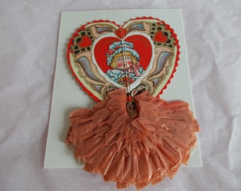 May include: Vintage Valentine's Day card with a heart-shaped design featuring a girl in a blue dress and a red heart. The card has a ruffled, peach-colored paper trim.