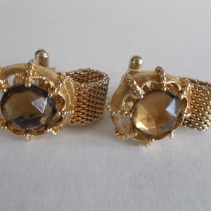 Pu&ograve; includere: Un paio di gemelli d'oro con una pietra di vetro sfaccettata marrone al centro di ogni gemello. I gemelli sono collegati da una catena d'oro.