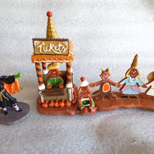 May include: A whimsical Halloween-themed figurine set. It features a witch in a black and orange outfit, a ticket booth with a gingerbread-like design, and several gingerbread people. The ticket booth has the word "Tickets" on it.