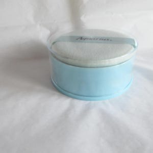 May include: A round, light blue powder compact with a clear lid. The compact contains a white powder puff and a light blue ribbon with the word "Aquarius" printed on it. The compact is sitting on a white surface.