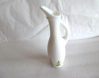 May include: A white ceramic pitcher with a handle and a spout. The pitcher has a slender body and a decorative green floral design near the base. The pitcher is likely used for pouring liquids.
