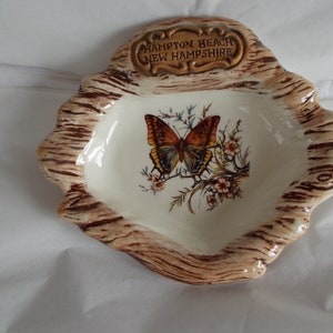 May include: A ceramic ashtray shaped like a tree stump with a brown and white woodgrain design. The ashtray features a painted butterfly with brown, orange, and blue wings. The text "Hampton Beach New Hampshire" is printed on a gold plaque on the top of the ashtray.
