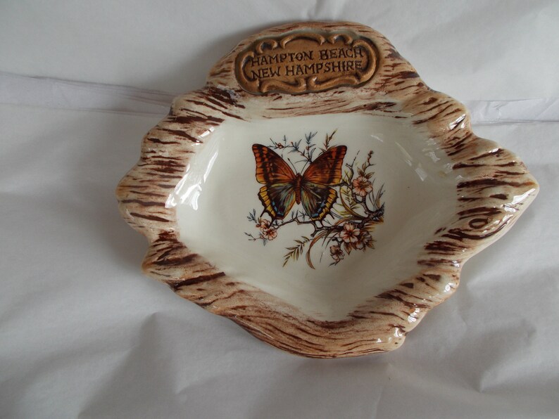 May include: A ceramic ashtray shaped like a tree stump with a brown and white woodgrain design. The ashtray features a painted butterfly with brown, orange, and blue wings. The text "Hampton Beach New Hampshire" is printed on a gold plaque on the top of the ashtray.