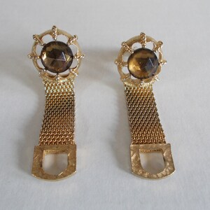 Pu&ograve; includere: Un paio di orecchini dorati con una pietra marrone sfaccettata incastonata in una cornice di metallo decorata. Gli orecchini sono attaccati a una lunga catena di metallo dorato.