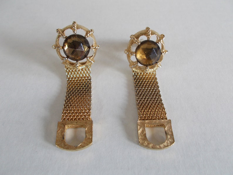 Pu&ograve; includere: Un paio di orecchini dorati con una pietra marrone sfaccettata incastonata in una cornice di metallo decorata. Gli orecchini sono attaccati a una lunga catena di metallo dorato.