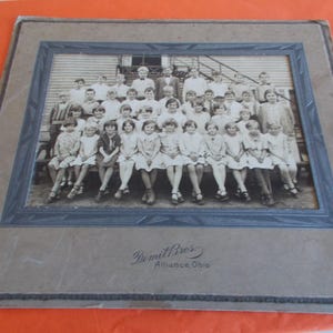 May include: A vintage black and white group photograph of school children mounted on a dark gray cardboard frame with a decorative border. The text Dimil Bros Alliance Ohio is printed in black ink at the bottom center of the mounting board.