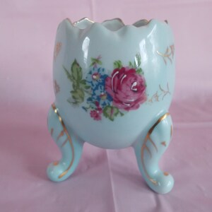 May include: A light blue ceramic egg-shaped planter with three gold-trimmed feet and a floral design. The planter features a pink rose and other flowers in shades of blue and white.