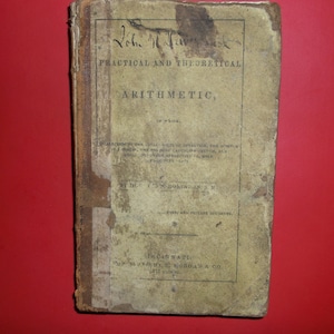 Puede incluir: Un libro antiguo titulado "Practical and Theoretical Arithmetic" de John M. Harwood. El libro está encuadernado en cuero marrón con una cubierta descolorida y amarillenta. El texto de la cubierta está en tinta negra.