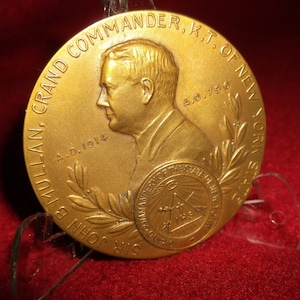 May include: A gold-coloured medallion with a profile of a man and the inscription "SIR JOHN B. MULLAN, GRAND COMMANDER, K.T. OF NEW YORK, A.D. 1914". The medallion also features a laurel wreath and the words "COMMANDERY OF THE STATE OF NEW YORK".