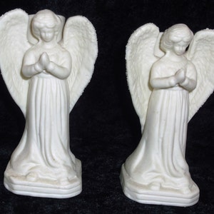 以下が含まれることがあります： 翼と祈りのポーズをした白い磁器製の天使のキャンドルホルダー2つ。天使は正方形の台座の上に立っています。