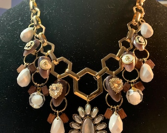 May include: A gold chain necklace with a variety of charms, including white and brown wooden beads, white and clear glass beads, and silver and gold metal charms. The necklace has a large, silver and gold metal centerpiece with a floral design.