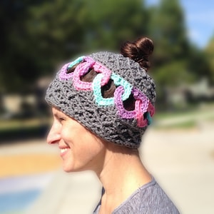 Puede incluir: Un gorro de ganchillo gris con un diseño de eslabones de cadena en rosa, turquesa y morado. El gorro tiene un patrón texturizado y está diseñado para acomodar un moño. El logo "Krazy Kabbage" es visible en la esquina inferior izquierda.