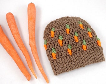 May include: A brown crocheted hat with orange and green carrot designs. The hat is sitting on a white surface with three orange carrots.