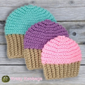 May include: Three crocheted hats in the shape of cupcakes. The hats are in teal, purple, and pink with a brown bottom. The hats are on a wooden surface.