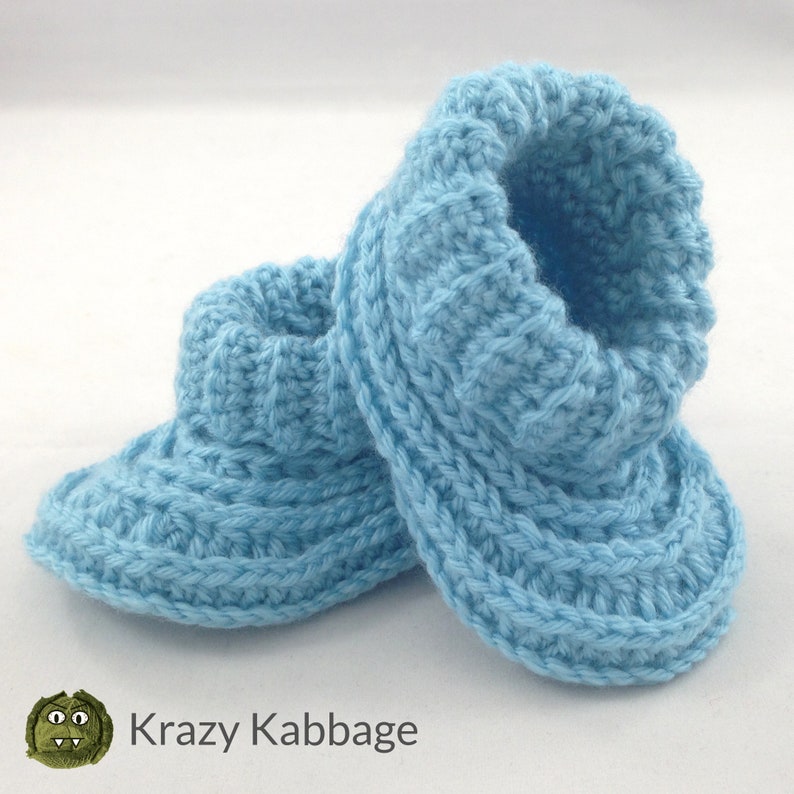 May include: A pair of light blue, hand-knitted baby booties. The booties are made of soft yarn and feature a ribbed cuff and a textured stitch pattern. The booties are designed to keep a baby's feet warm and cozy. The logo "Krazy Kabbage" is in the bottom left corner.