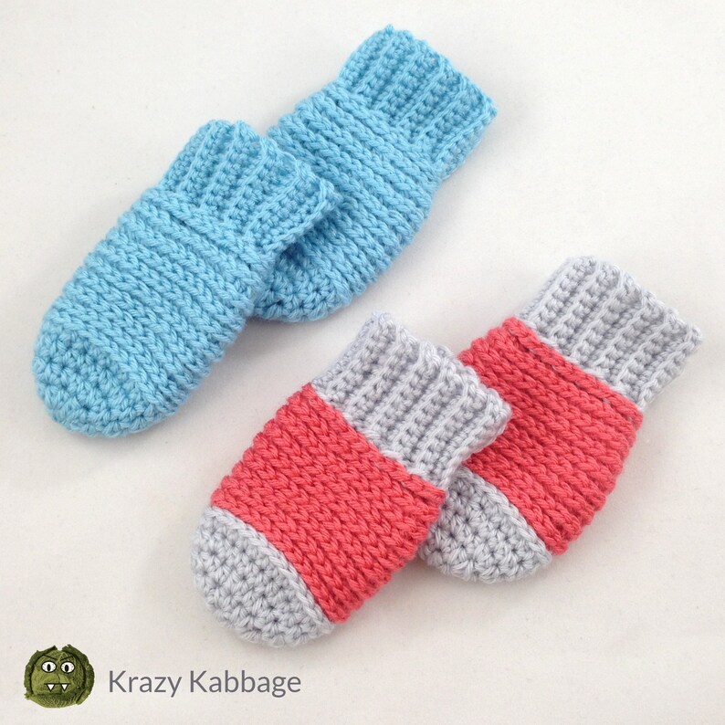 May include: Two pairs of handmade crochet baby booties. One pair is light blue, and the other is gray with a coral band. The booties are made with a ribbed stitch and have a soft, textured appearance. The logo "Krazy Kabbage" is in the bottom left corner.