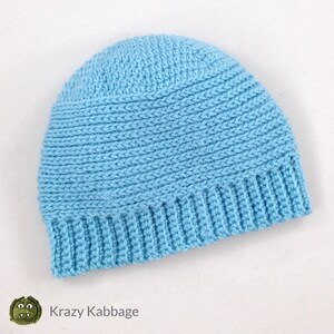 May include: A light blue crochet beanie hat. The hat features a ribbed brim and a textured stitch pattern on the crown. The hat is handmade and has the text "Krazy Kabbage" on the bottom left corner.