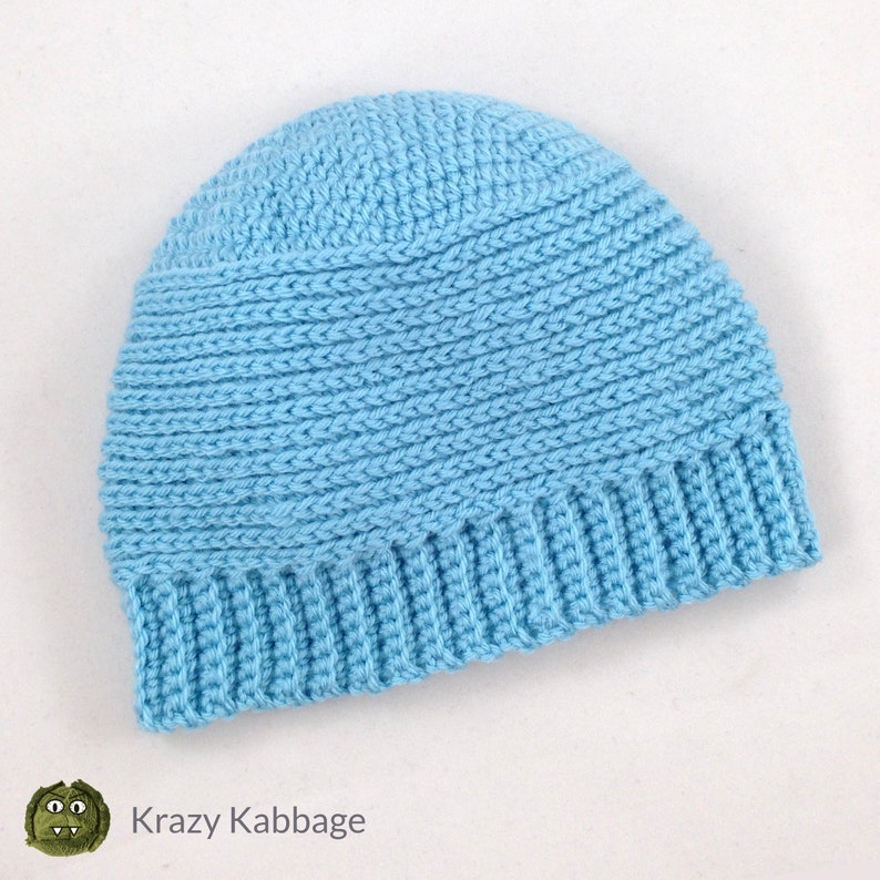 May include: A light blue crochet beanie hat. The hat features a ribbed brim and a textured stitch pattern on the crown. The hat is handmade and has the text "Krazy Kabbage" on the bottom left corner.