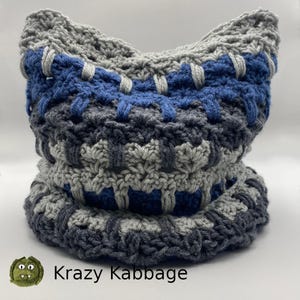 May include: A gray and blue crocheted cowl with a textured pattern. The cowl is folded in half and the bottom edge is visible. The text "Krazy Kabbage" is visible at the bottom of the image.