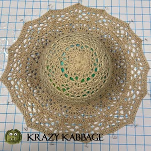 May include: A beige crochet hat with a lacy trim is pinned to a gridded surface. The hat is being prepared for sewing. The text "KRAZY KABBAGE" is visible at the bottom of the image.