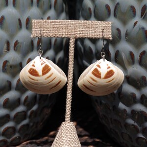 May include: A pair of seashell earrings displayed on a burlap-covered stand. The earrings are cream-colored with brown markings and silver-colored hooks. The background features a textured, blue and brown ceramic vase.