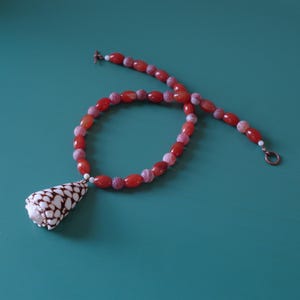 May include: A red agate bead necklace with a shell pendant. The necklace is made of oval-shaped beads with a white and red pattern. The shell pendant is a cone shape with a brown and white pattern.
