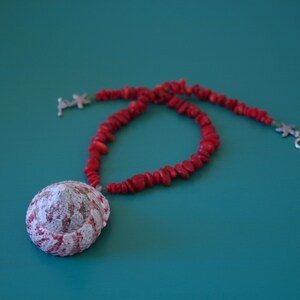 May include: A red coral necklace with a large white seashell pendant. The necklace is on a teal background.