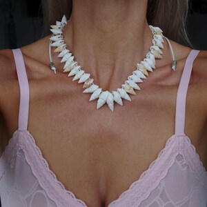 May include: A white seashell necklace with a natural, organic design. The necklace features a series of pointed, cone-shaped shells strung together with a white cord. The shells are primarily white with some tan accents.