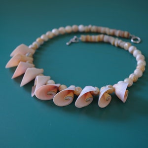 May include: A necklace made of small, white, shell beads with a cluster of larger, pink, shell pendants. The necklace is on a teal surface.