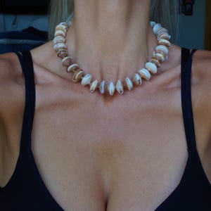 May include: A close-up of a woman wearing a black tank top with a shell necklace. The necklace is made of natural shells in shades of brown and white.