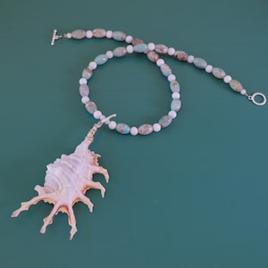 May include: A white and blue gemstone necklace with a seashell pendant. The necklace is made of oval-shaped beads and has a silver clasp.