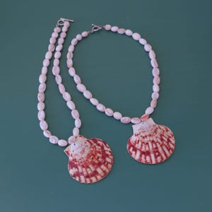 May include: Two necklaces with white and pink oval beads and a large, red and white striped seashell pendant. The necklaces have a silver clasp. The background is a solid teal color.