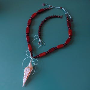 May include: A red wooden bead necklace with a seashell pendant and a smaller red wooden bead bracelet. The necklace and bracelet are tied together with a light blue ribbon.