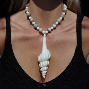 May include: A white shell pendant necklace with a long, spiraled shell charm. The necklace is made of white and gray beads.