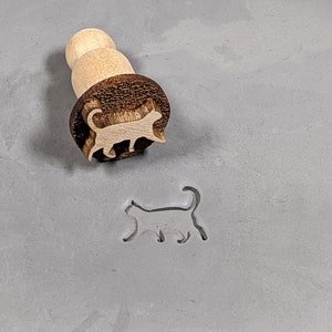 May include: A wooden cat-shaped cookie cutter with a brown handle. The cutter is used to create a cat-shaped impression in dough.
