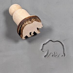 May include: A wooden bear stamp with a brown handle. The stamp is used to create an imprint of a bear on clay or dough.