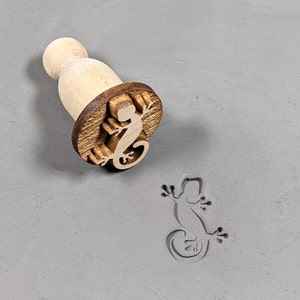 May include: A wooden stamp with a carved gecko design. The stamp is used to create an impression of a gecko on a surface.