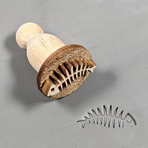 May include: A wooden stamp with a fish bone design. The stamp is used to create an imprint of the fish bone on clay or dough.