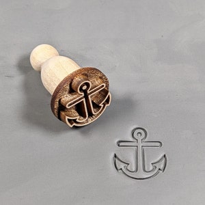 May include: A wooden stamp with a carved anchor design. The stamp is used to create an imprint of the anchor on a surface.
