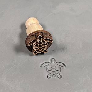 May include: A wooden stamp with a detailed carving of a sea turtle. The stamp is used to create impressions in clay or dough.