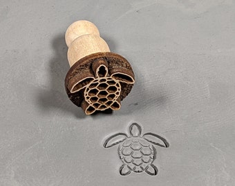 May include: A wooden stamp with a detailed carving of a sea turtle. The stamp is used to create impressions in clay or dough.
