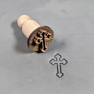 May include: A wooden stamp with a cross design. The stamp is used to create an impression of a cross in clay or dough.