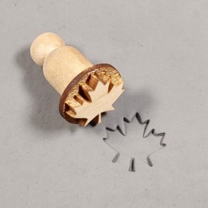 May include: A wooden maple leaf stamp with a brown handle. The stamp is used to create a maple leaf impression in clay or dough.