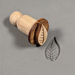 May include: A wooden leaf-shaped stamp with a brown handle. The stamp is used to create impressions in clay or dough.