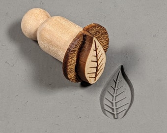 May include: A wooden leaf-shaped stamp with a brown handle. The stamp is used to create impressions in clay or dough.