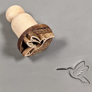 May include: Wooden stamp with a hummingbird design. The stamp is made of light brown wood and has a dark brown handle. The hummingbird is carved into the wood and is facing to the right. The stamp is on a gray surface.