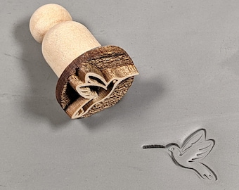 May include: Wooden stamp featuring a hummingbird design. The stamp is crafted from light brown wood and has a dark brown handle. The hummingbird is carved into the wood and is facing to the right. The stamp is on a grey surface.