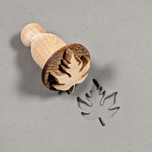 May include: A wooden stamp with a maple leaf design. The stamp is used to create an imprint of the leaf on a surface.