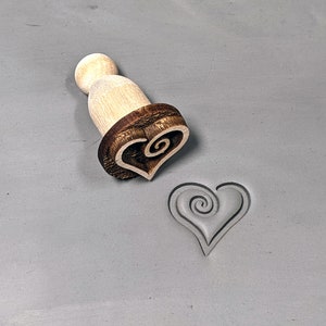May include: A wooden heart-shaped stamp with a spiral design. The stamp is used to create an impression on clay or dough.